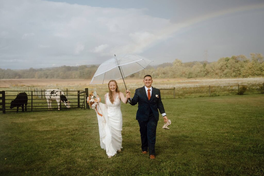 a bride and groom walking through the Columbus Ohio area countryside under an umbrella and a rainbow