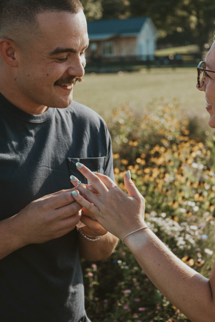 A fiancé slipping an engagement ring onto their partner’s finger in a wildflower meadow.