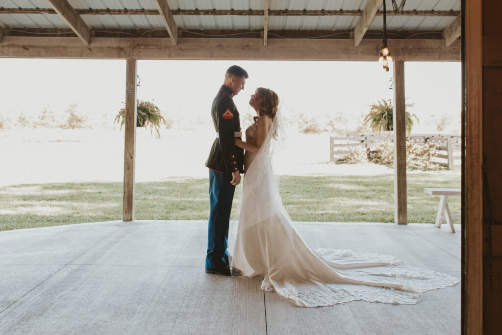 Newlyweds stand close together beneath a rustic pavilion at 22 Acres Farm, just outside Columbus, Ohio. The groom wears a Marine Corps dress uniform, and the bride's flowing veil and train create a timeless, elegant scene.