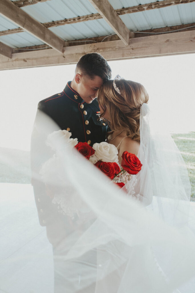 A Marine groom and his bride share an intimate moment at 22 Acres Farm near Columbus, Ohio. The bride’s veil softly drapes around them as they lean in, holding a bouquet of red and white roses.