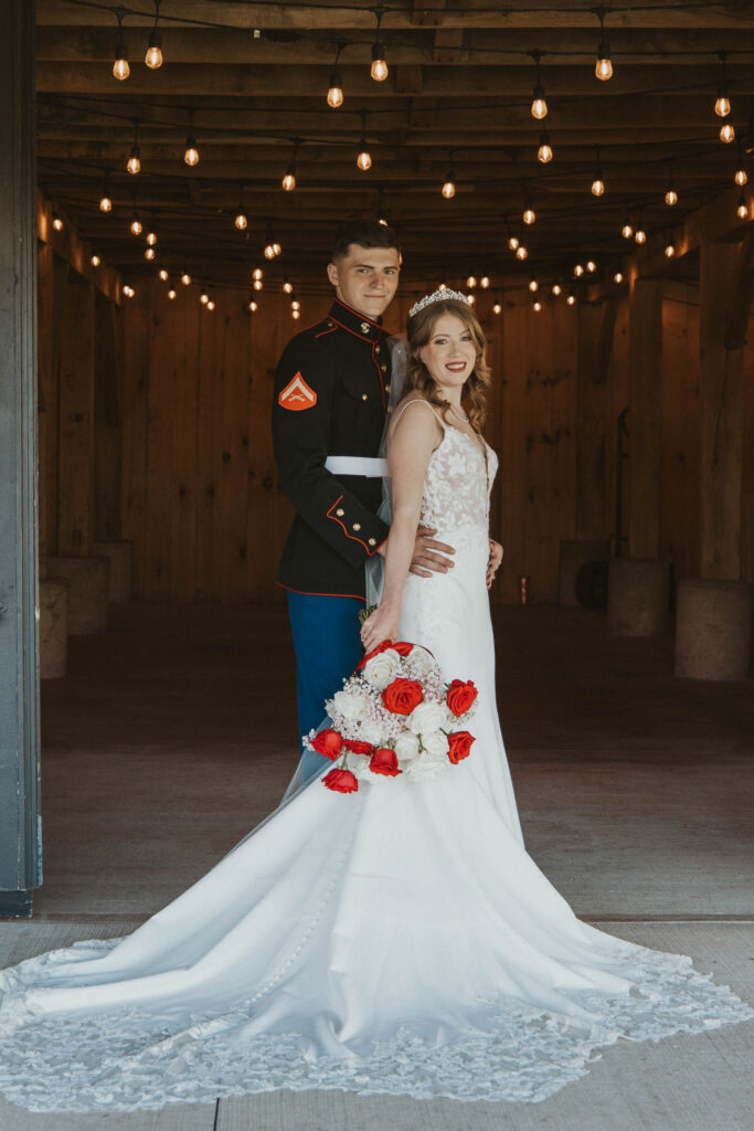 A Marine groom and his bride pose under twinkling lights at 22 Acres Farm near Columbus, Ohio. The bride’s gown cascades behind her as she holds a bouquet of red and white roses.