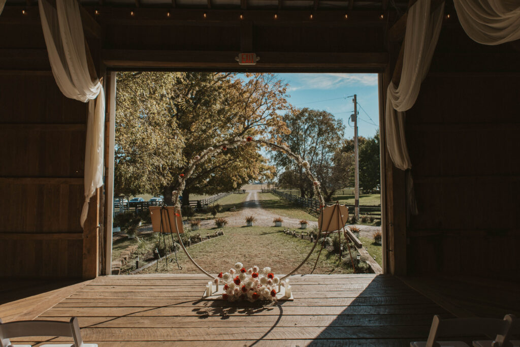 The view from inside a barn wedding venue at 22 Acres Farm, just outside Columbus, Ohio, looking out onto a picturesque country road. A circular floral arch, soft candlelight, and draped fabric add a romantic touch to the rustic setting.