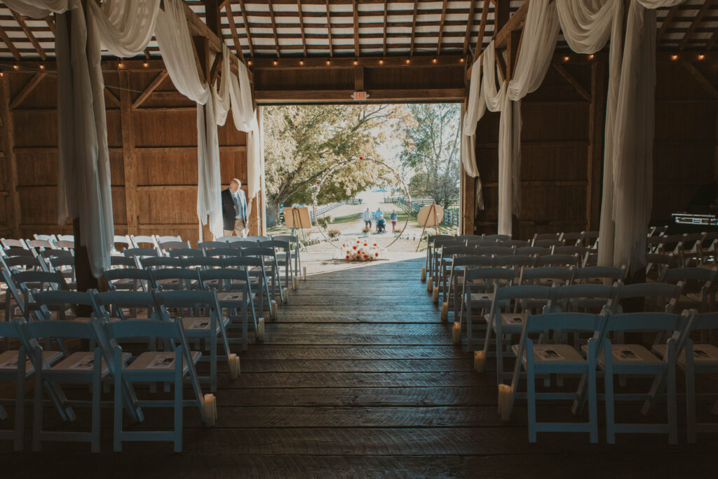 A beautifully decorated barn ceremony setup at 22 Acres Farm near Columbus, Ohio. Rows of white chairs line the wooden aisle, leading to a circular floral arch framing the sunlit countryside beyond. Soft drapery and candlelight enhance the romantic, rustic charm.