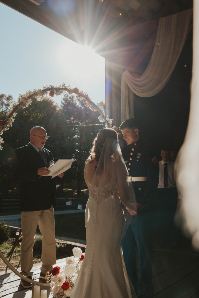 A bride and her Marine groom stand hand in hand during their wedding ceremony at 22 Acres Farm near Columbus, Ohio. Sunlight streams through the open barn doors, casting a warm glow over the moment.