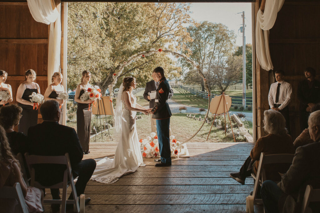 A bride and her Marine groom hold hands during their wedding ceremony at 22 Acres Farm near Columbus, Ohio. Sunlight streams through the open barn doors, illuminating the couple beneath a floral circular arch.