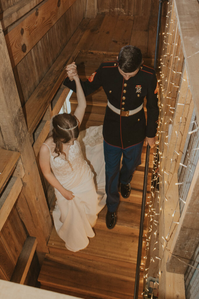 A Marine groom leads his bride down a wooden staircase at 22 Acres Farm near Columbus, Ohio. Twinkling string lights and rustic barn walls frame the intimate moment.