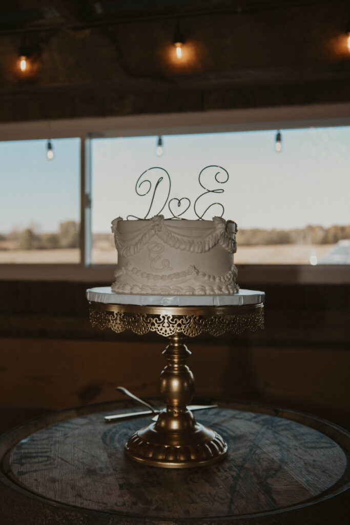 A white wedding cake with intricate piping and a custom 'D & E' topper sits on a gold stand at 22 Acres Farm near Columbus, Ohio. Warm string lights and a rustic barn window frame the scene.