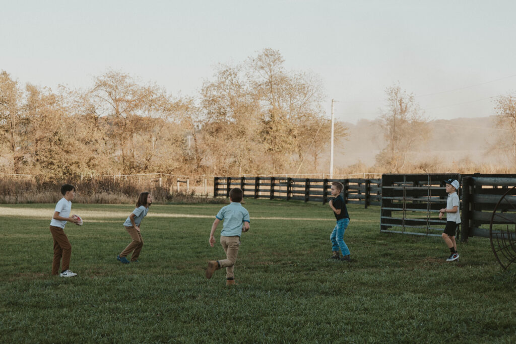 Children play a game of football in an open field at 22 Acres Farm near Columbus, Ohio. The rustic wooden fences and autumn trees in the background add to the charming countryside setting.