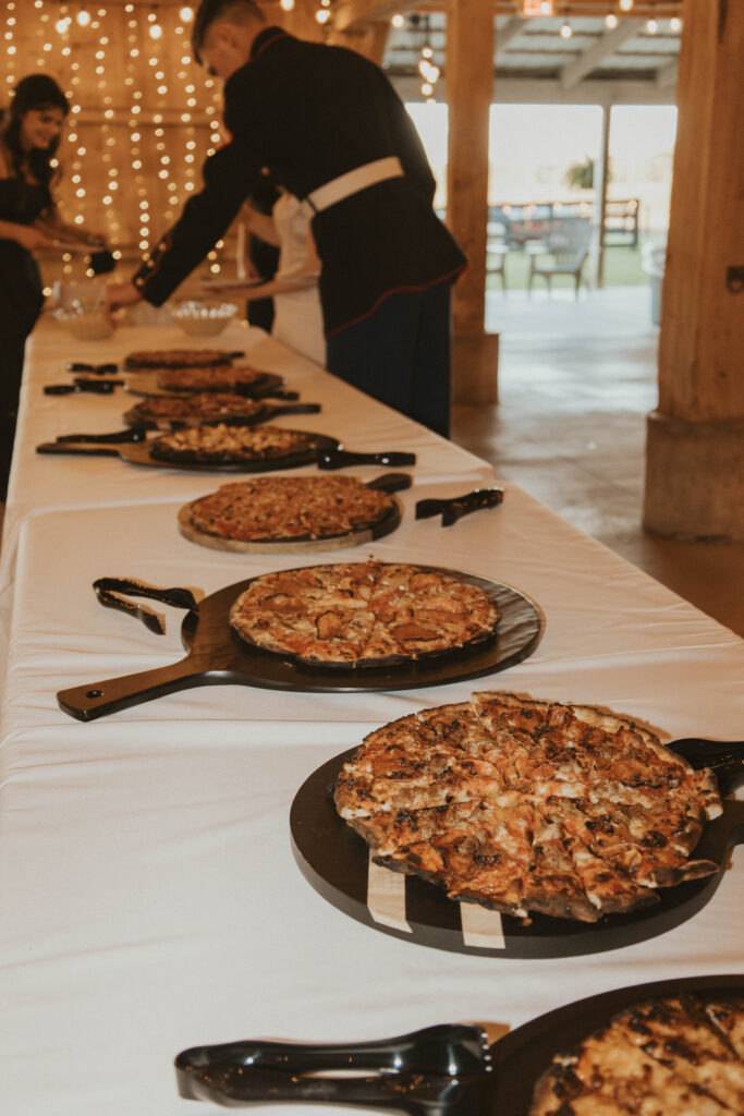 A wedding reception at 22 Acres Farm near Columbus, Ohio, featuring a rustic pizza buffet. The groom, dressed in Marine blues, serves himself as string lights glow in the background.