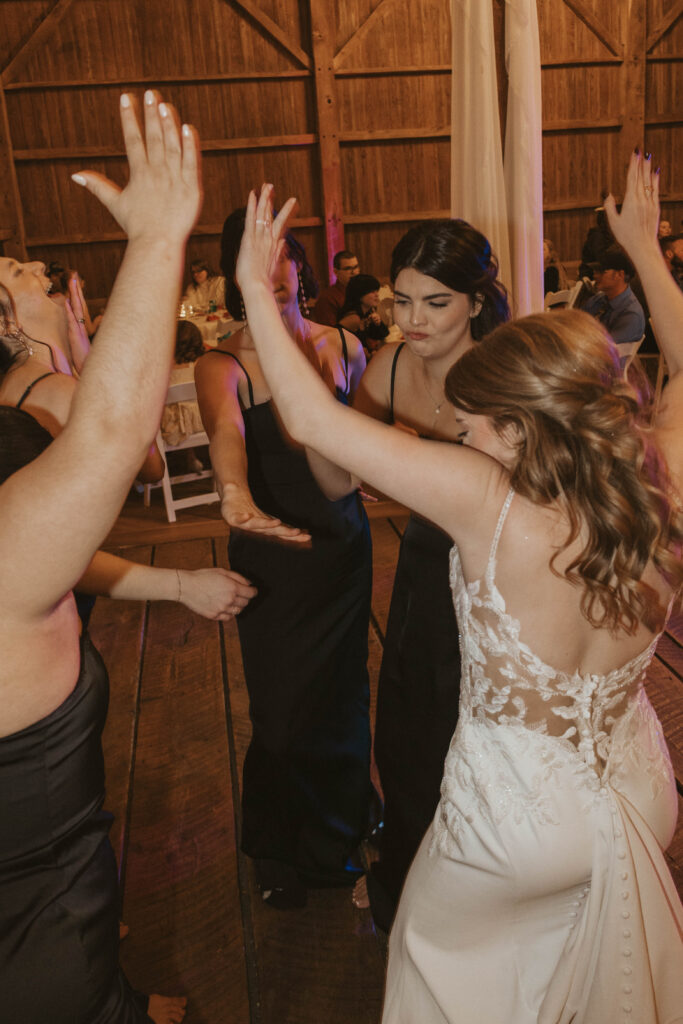 The bride dances with friends at her wedding reception in the barn at 22 Acres Farm near Columbus, Ohio. Laughter and raised hands fill the rustic wooden space.