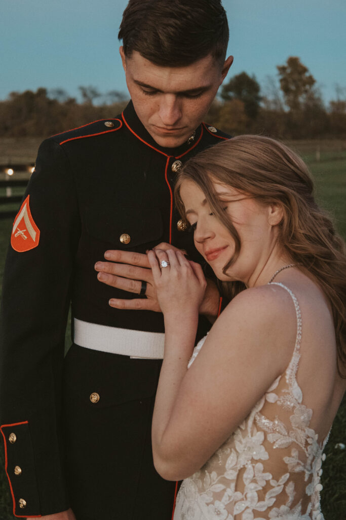 A close-up of a Marine groom and his bride at 22 Acres Farm near Columbus, Ohio. She holds his hand over his chest, her wedding ring visible as she smiles softly in the fading sunlight.