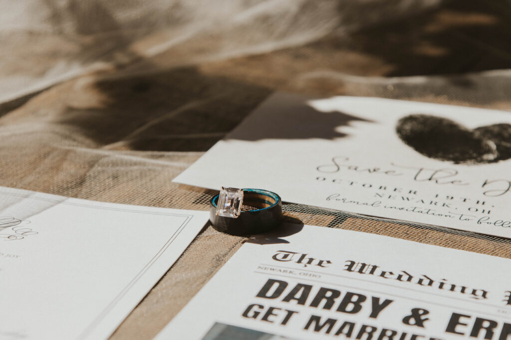 Wedding details from a 22 Acres Farm wedding near Columbus, Ohio. The rings rest on invitations and a newspaper-style wedding announcement, all set against rustic burlap.