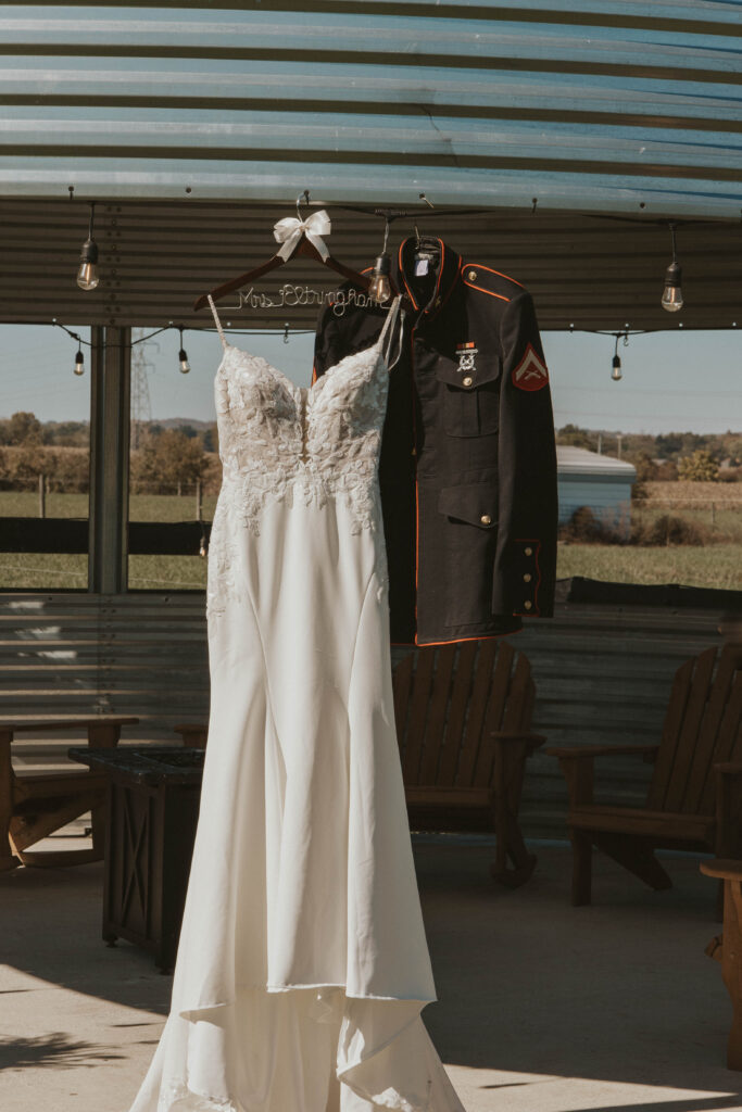 A bridal gown and a Marine groom’s uniform hang side by side in the Grain Bin Gazebo at 22 Acres Farm near Columbus, Ohio. The open-air structure, adorned with string lights and wooden seating, captures the farm’s rustic elegance.