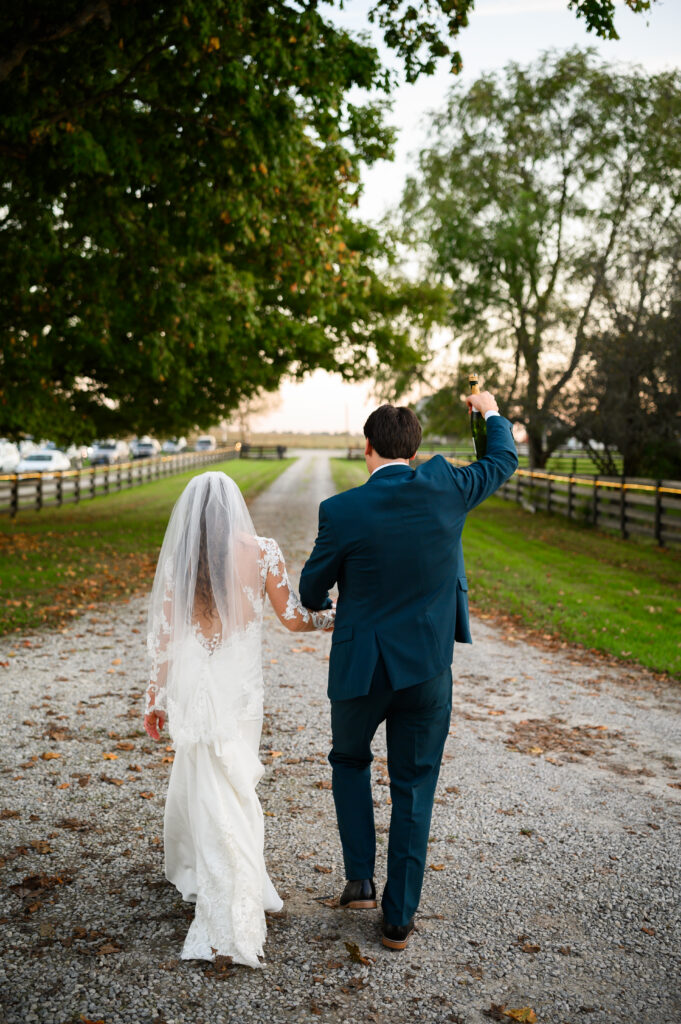 The couple walks hand-in-hand down a gravel farm lane, with the groom holding a champagne bottle triumphantly. The tree-lined path at 22 Acres Farm frames this carefree exit from their outdoor Ohio wedding ceremony.