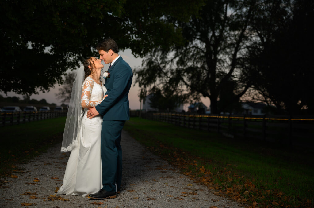 A bride and groom embrace beneath a canopy of trees at sunset during their outdoor Ohio wedding at 22 Acres Farm. Fallen leaves and soft light set a romantic tone on the venue’s scenic gravel lane.