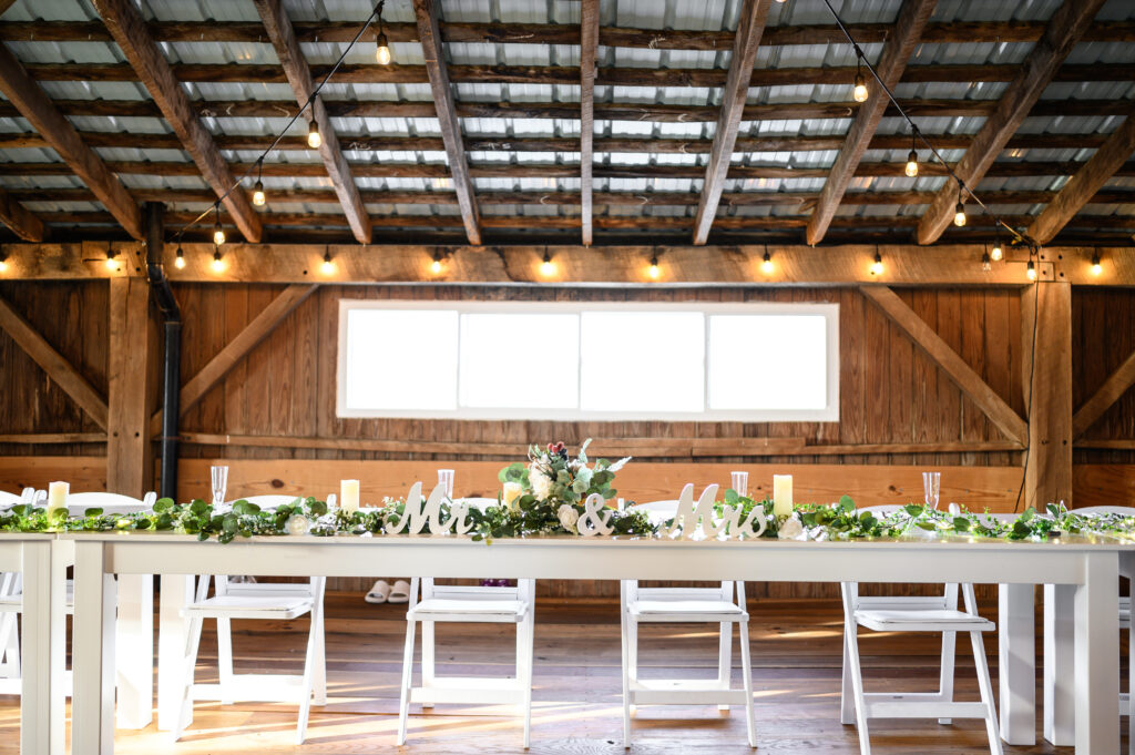 A white sweetheart table at 22 Acres Farm is decorated with eucalyptus garland, candles, and a “Mr. & Mrs.” sign. This cozy setup highlights the couple’s rustic barn reception after their outdoor Ohio wedding.